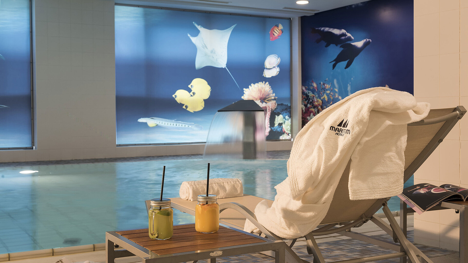 Pool Relaxation area by the pool at the Maritim proArte Hotel Berlin with a lounger, towel, and fresh juices next to a waterfall.