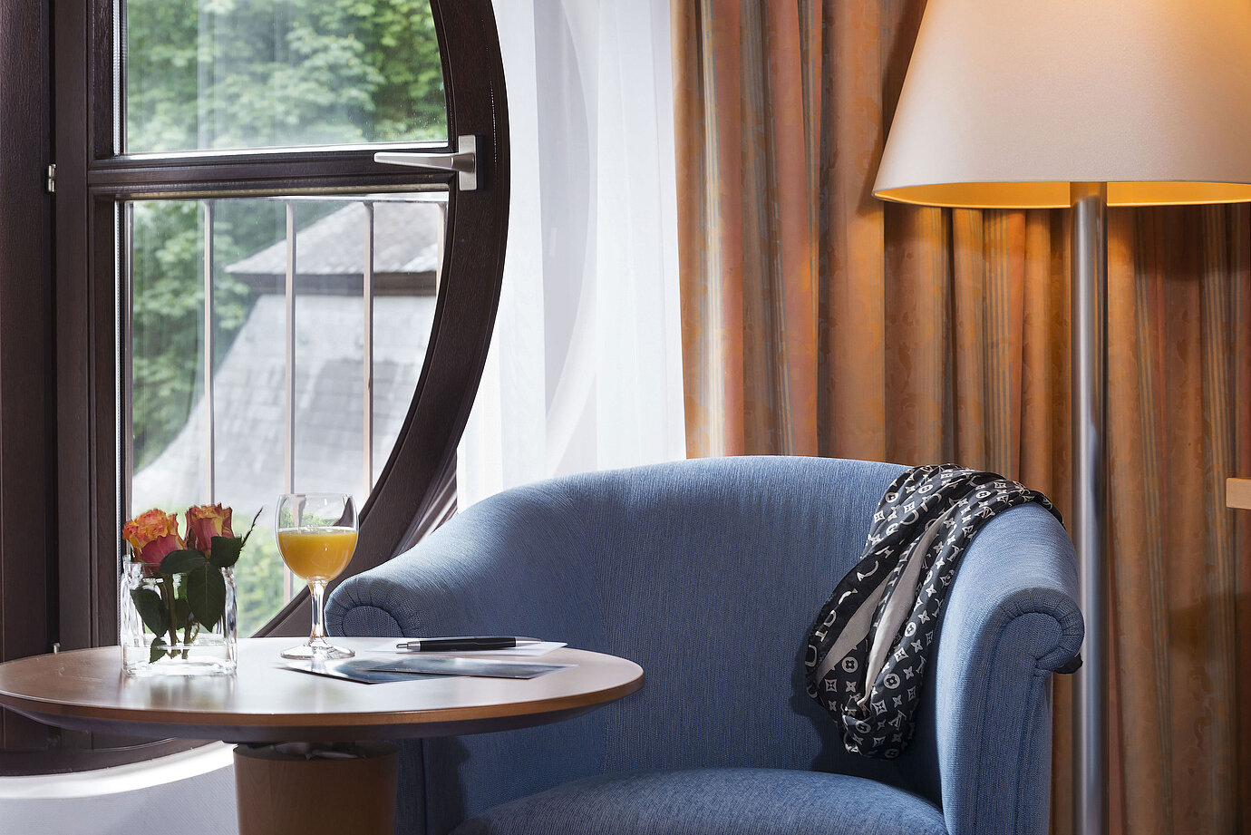 Acogedora zona de descanso en la Habitación Classic del Maritim Fulda con sillón azul,ventana redonda,flores y zumo de naranja