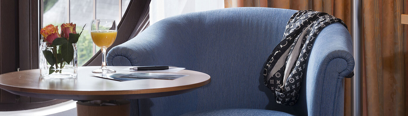 Coin salon confortable dans la chambre Classic du Maritim Hotel Fulda avec fauteuil bleu,fenêtre ronde,fleurs et jus d’orange
