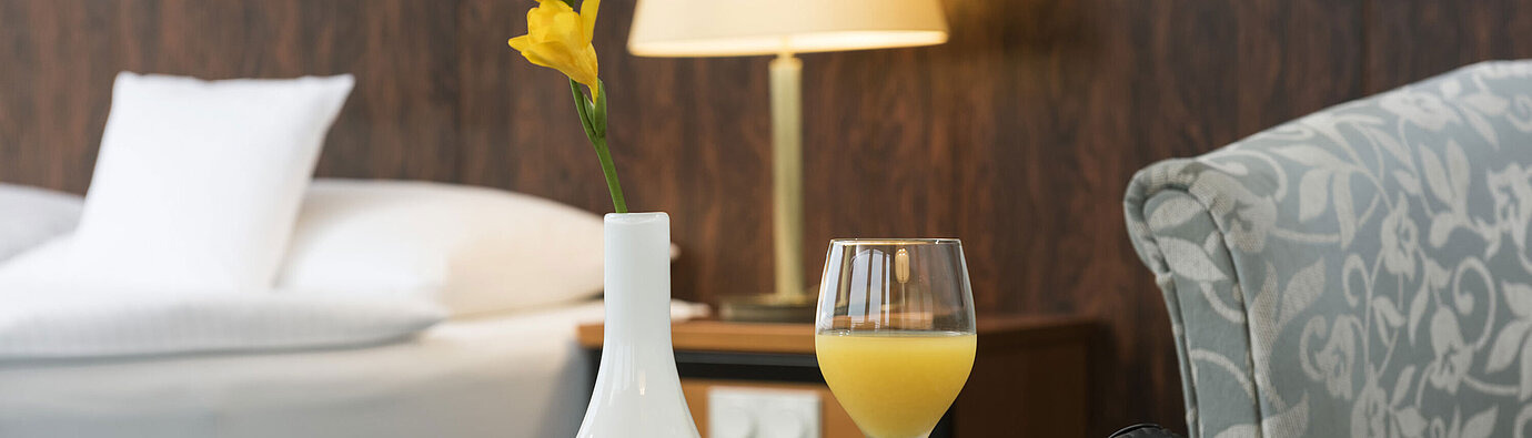 Detalle de la sala Detalles de una habitación del Maritim Hotel Kiel: vaso de zumo de naranja, auriculares y jarrón sobre la mesa, una cama al fondo.