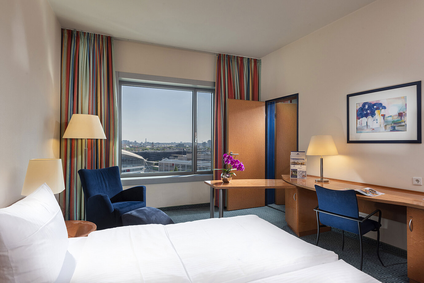 Camera familiare Comfort del Maritim Hotel Frankfurt con letto matrimoniale, scrivania, tende colorate e vista città.