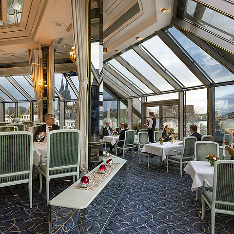 Restaurante elegante con techo de vidrio y vistas a la catedral de Colonia y al puente Hohenzollern, comensales disfrutando la comida.