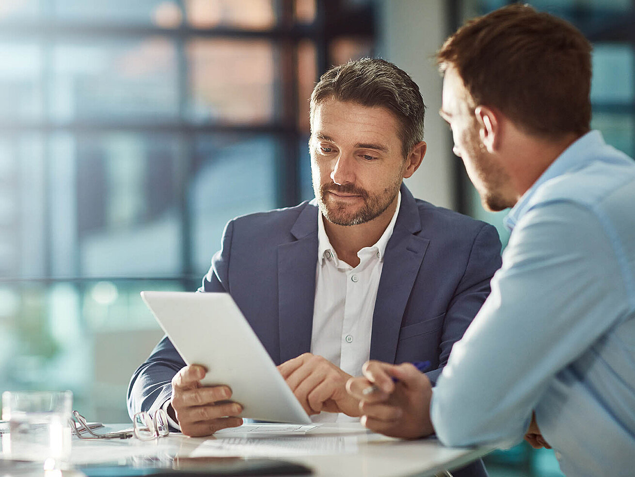 Two businessmen in the office discuss documents on a tablet.