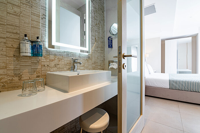 Modern bathroom in the Junior Suite at Maritim Antonine Hotel & Spa Malta featuring an illuminated mirror, stylish washbasin and a view into the bright sleeping area.