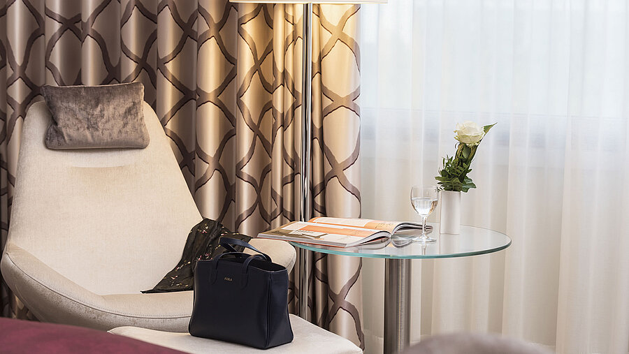 Détail d'une chambre Classic à l'hôtel Maritim Bonn avec un fauteuil confortable, rideau, table, verre, rose et sac à main. 