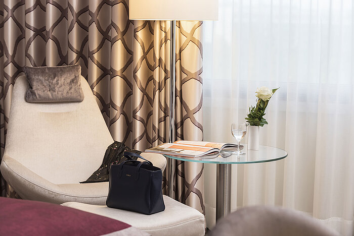 Dettaglio di una camera Classic al Maritim Hotel Bonn con poltrona comoda, tenda, tavolo, bicchiere, rosa e borsa. 