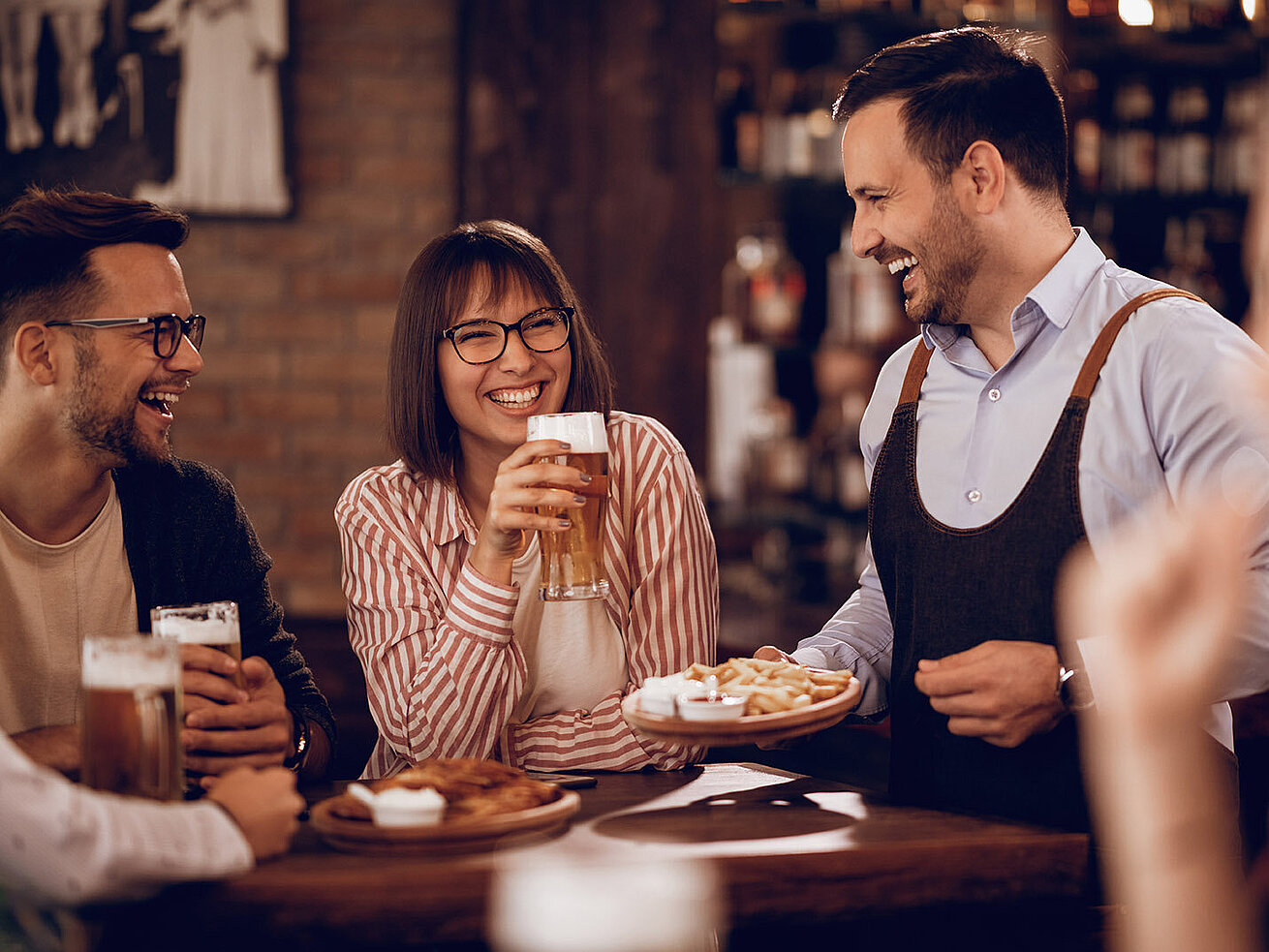 Smiling guests enjoy beer and snacks, served by friendly waiter in bar