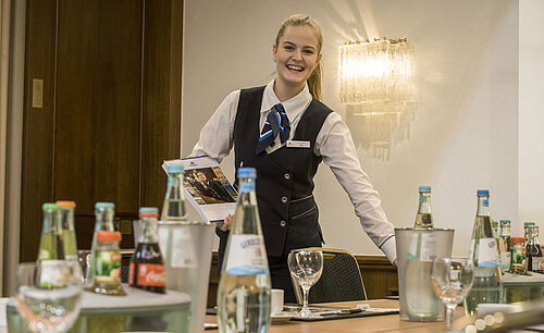 Aimable employée de service dans le salon Lenné du Maritim Hotel Bonn avec des boissons et des documents de conférence sur la table.