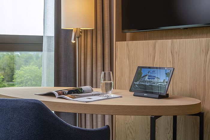 Escritorio en el Maritim Hotel Ingolstadt con tablet, revista y vaso de agua, con vistas a la naturaleza.