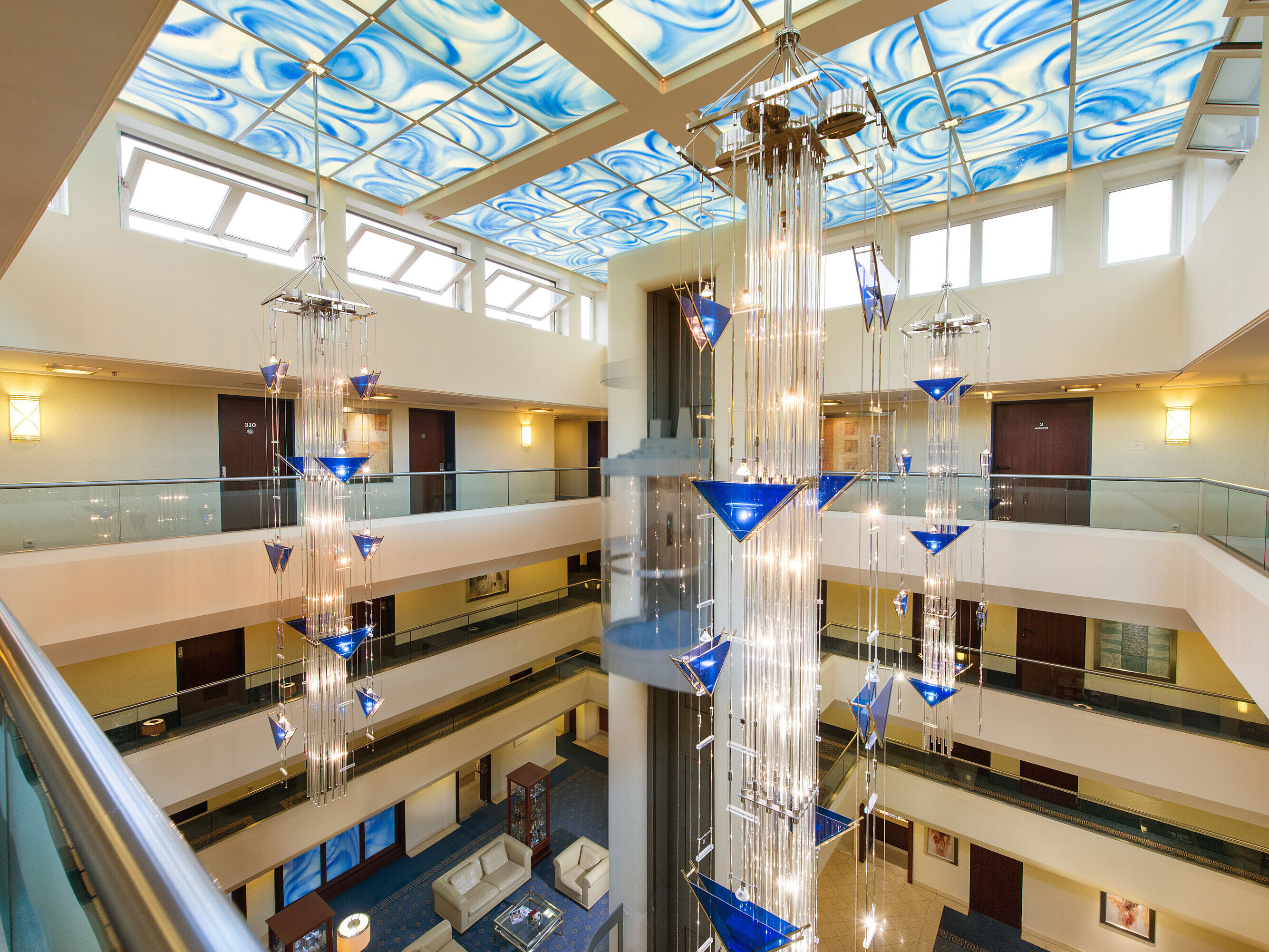 Atrio moderno en Maritim Hotel Fulda con ascensor de cristal, techo azul y lámpara artística.