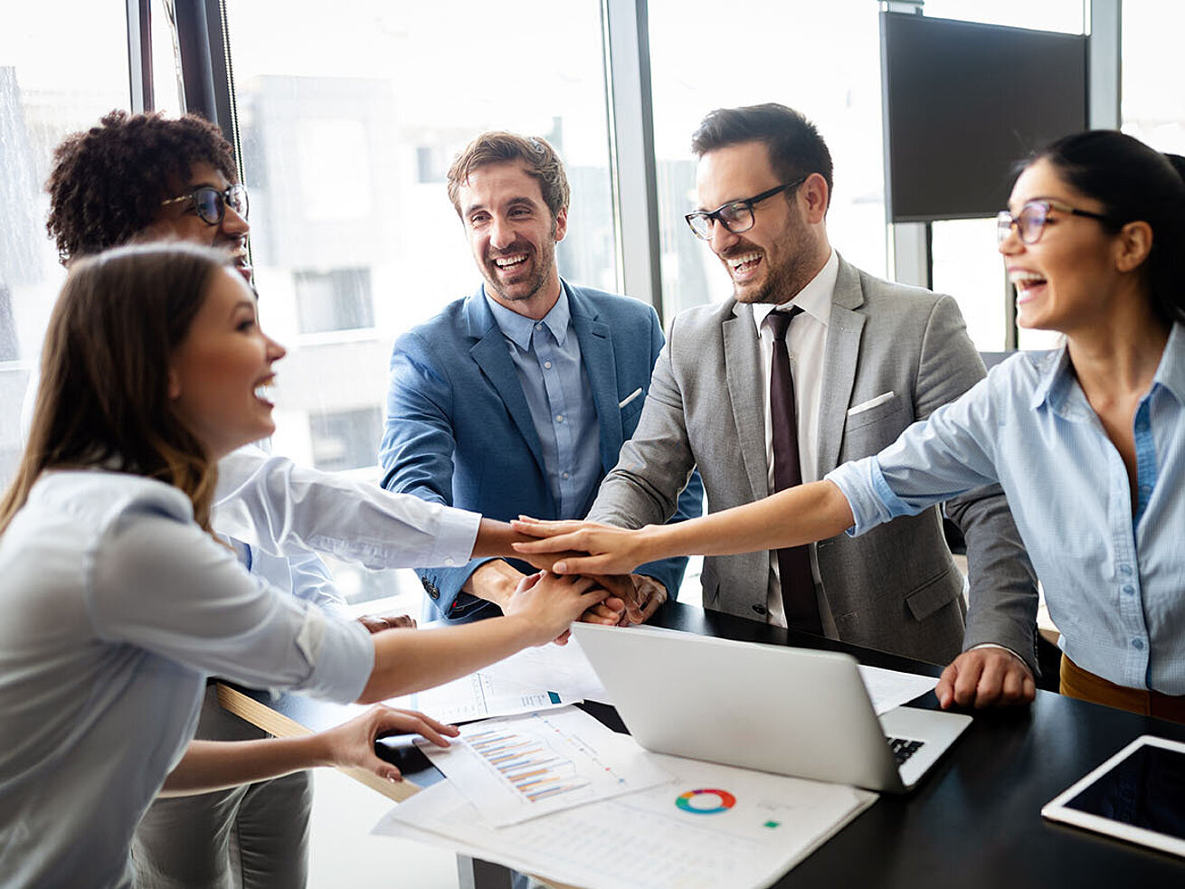 Motivated business team in office joining hands during meeting with laptop and documents on conference table