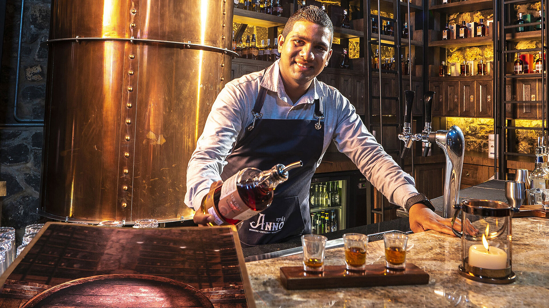 "The Distillery" Bartender serving spirits at the bar with copper still and illuminated shelves