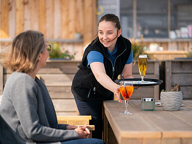 Une serveuse sert de l'Aperol Spritz et de la bière à un client sur la terrasse du Beach Lounge,avec la plage en arrière-plan