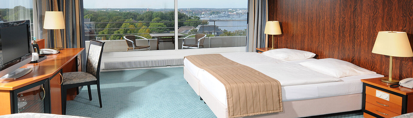 Chambre Comfort Chambre d’hôtel spacieuse avec lit double, coin salon et vue sur Kiel à travers la grande baie vitrée.