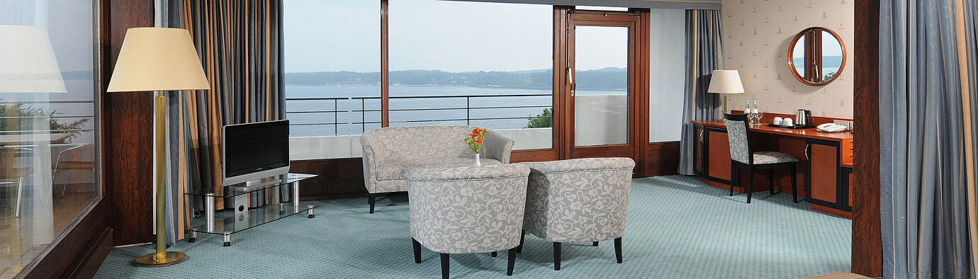 Junior Suite en el Maritim Hotel Kiel, con zona de estar, escritorio, TV y amplia ventana frontal con vistas al mar.