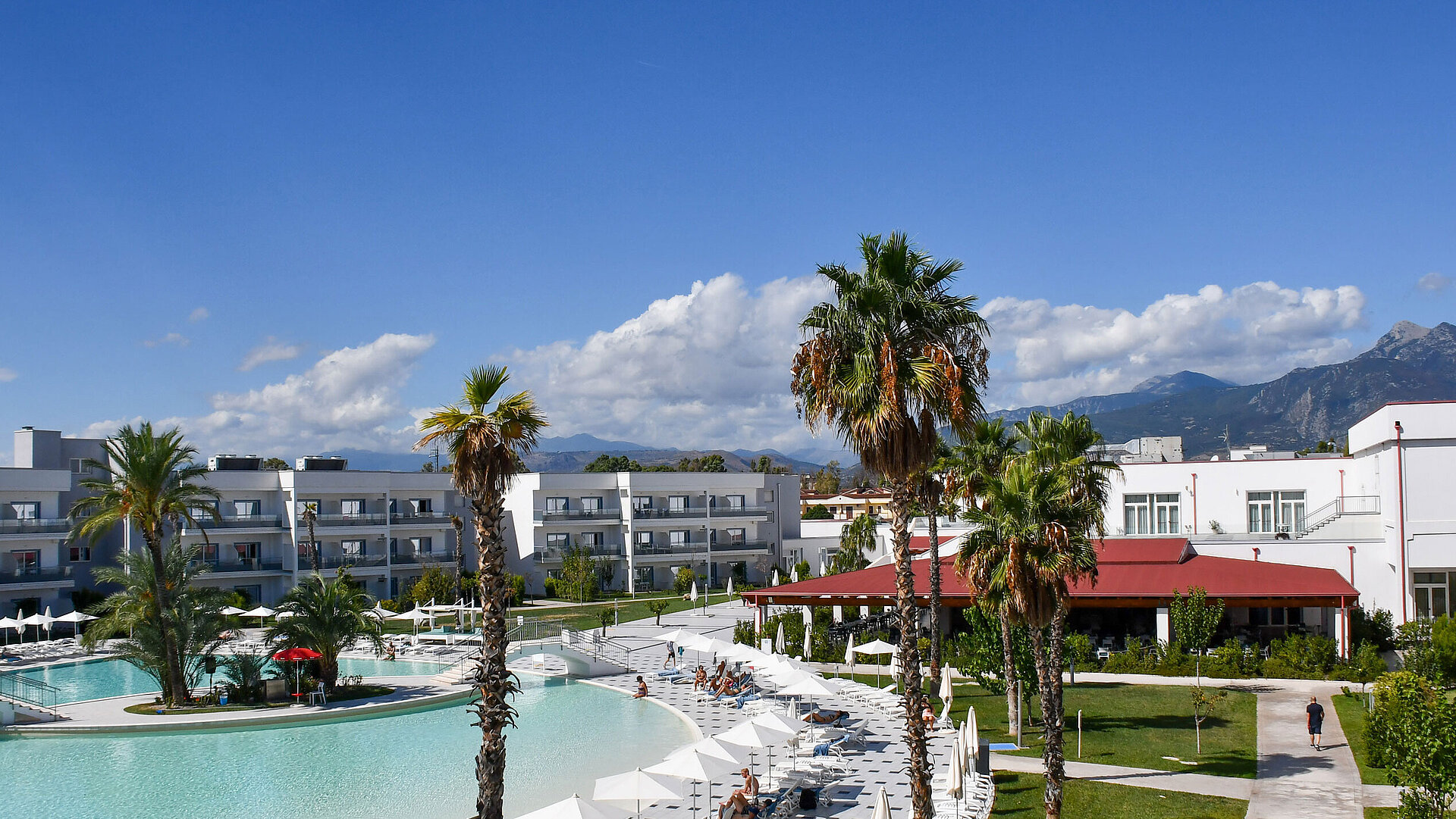 Panorama du Maritim Resort Calabria avec piscine, palmiers, bâtiments blancs et montagnes en arrière-plan sous un ciel clair.