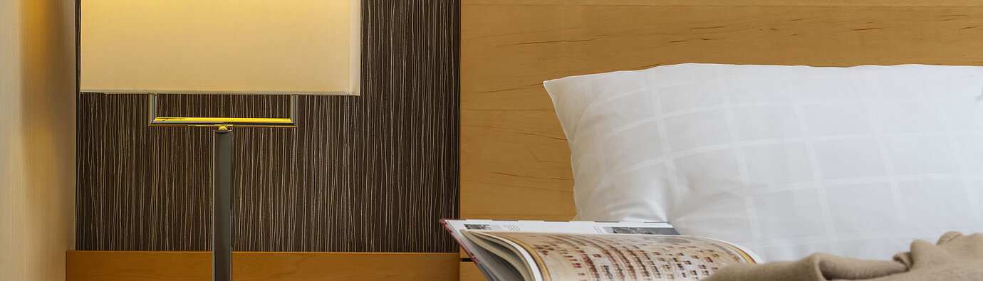 Primer plano de una mesita de noche en el Maritim Hotel Düsseldorf con lámpara, vaso de agua y libro en la cama.