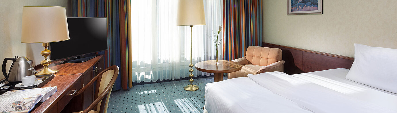 Chambre ComfortSMART Chambre Comfort lumineuse à l'Hôtel Maritim Cologne avec lit simple, bureau, TV, fauteuil et grandes fenêtres avec rideaux.