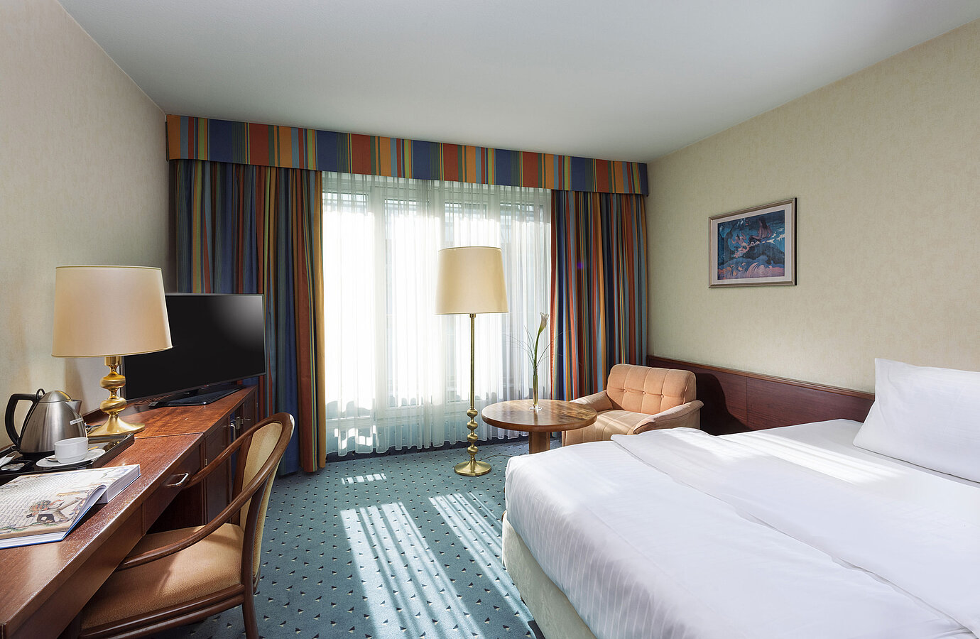 Camera Comfort luminosa al Maritim Hotel Colonia con letto singolo, scrivania, TV, poltrona e grandi finestre con tende.
