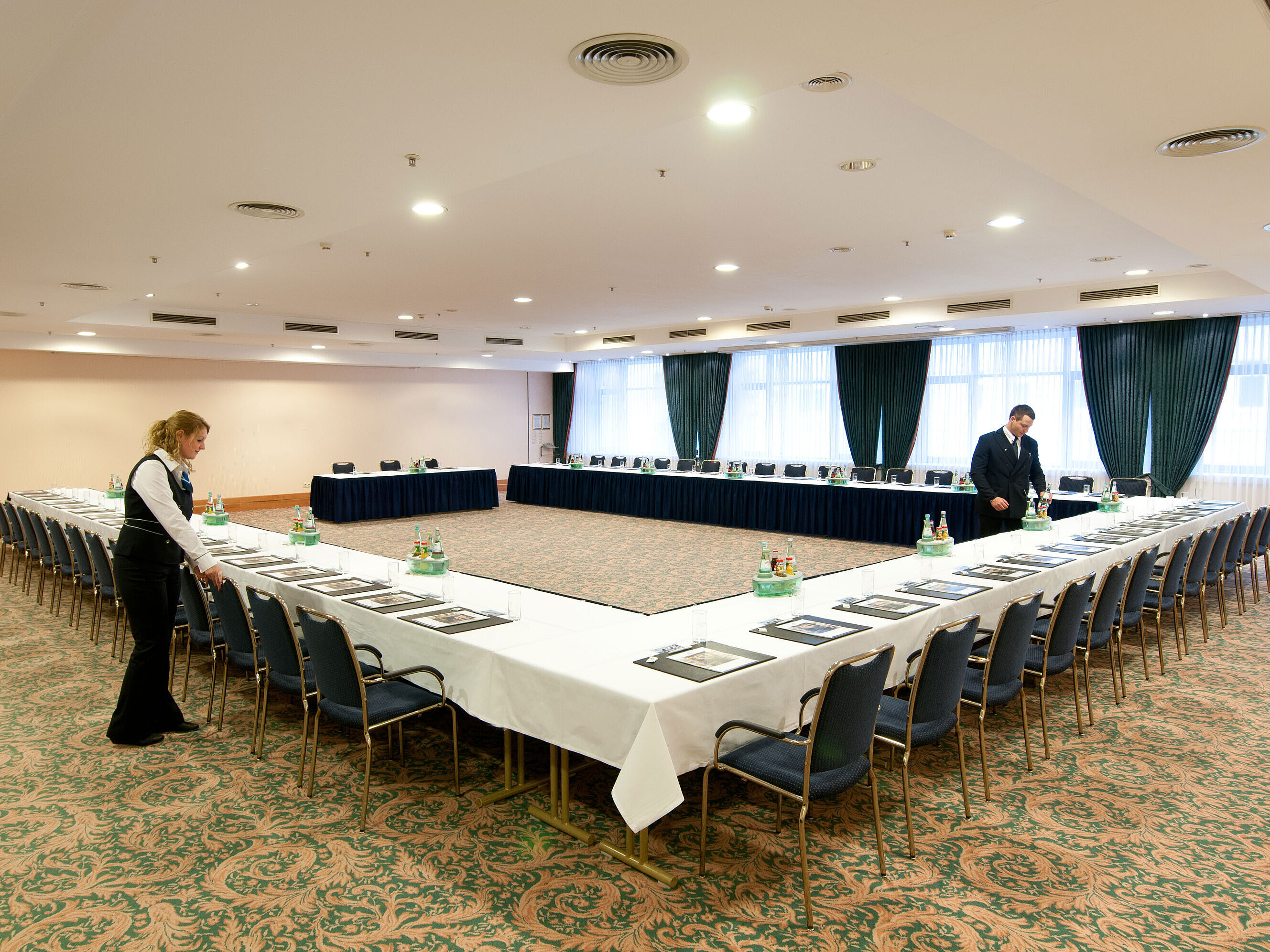 Sala de reuniones en el Maritim Hotel Magdeburg con asientos en forma de U, mesas listas y personal organizando.