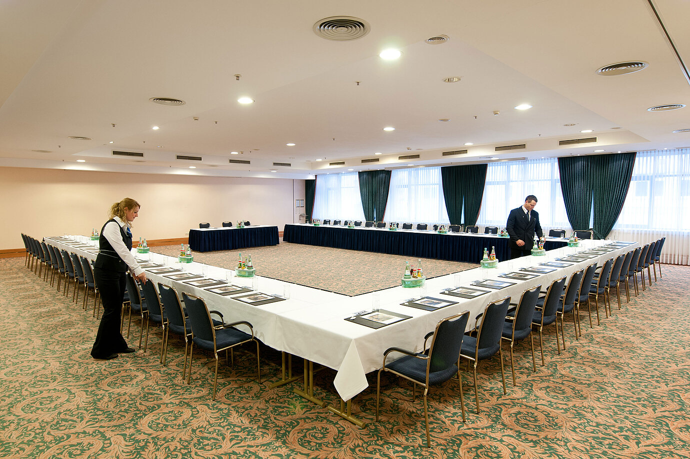 Sala de reuniones en el Maritim Hotel Magdeburg con asientos en forma de U, mesas listas y personal organizando.