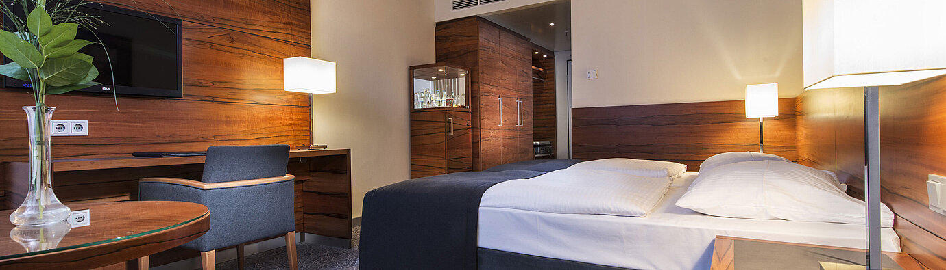 Camera d'albergo arredata presso il Maritim Hotel Monaco con letto confortevole, area lavoro, TV e pannelli in legno.