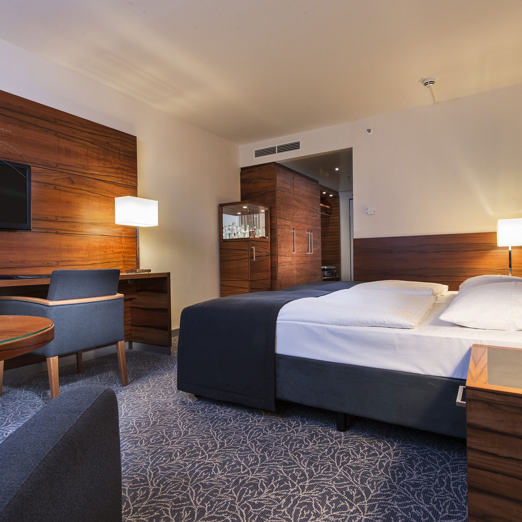 Camera Comfort Camera d'albergo arredata presso il Maritim Hotel Monaco con letto confortevole, area lavoro, TV e pannelli in legno.