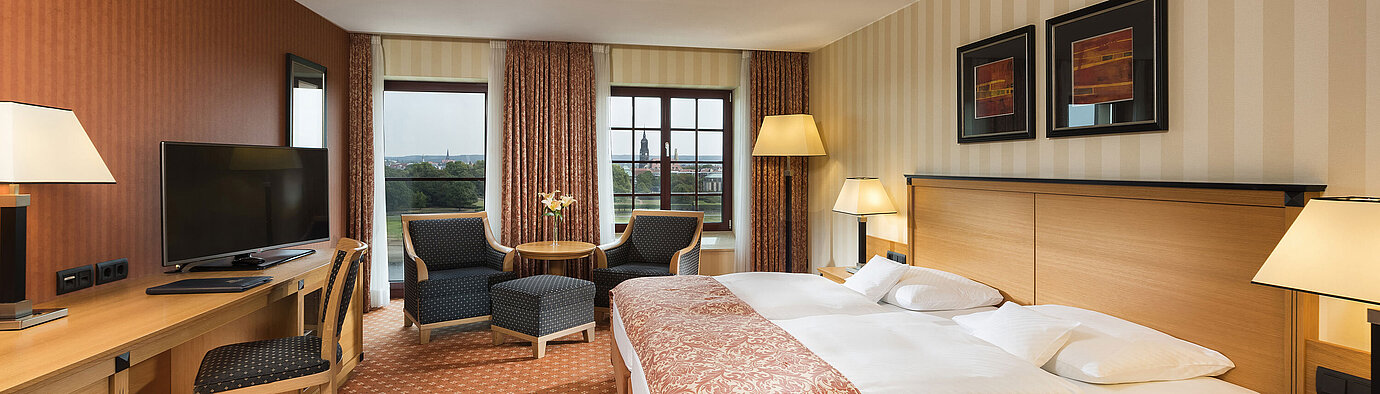 Camera superior al Maritim Hotel Dresden con letto matrimoniale elegante, scrivania e area salotto con vista sulla città.