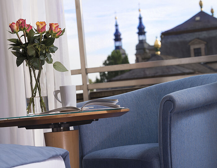  Detalle elegante de una habitación del Maritim Hotel Fulda con asiento azul, rosas y vistas a las torres barrocas de Fulda.