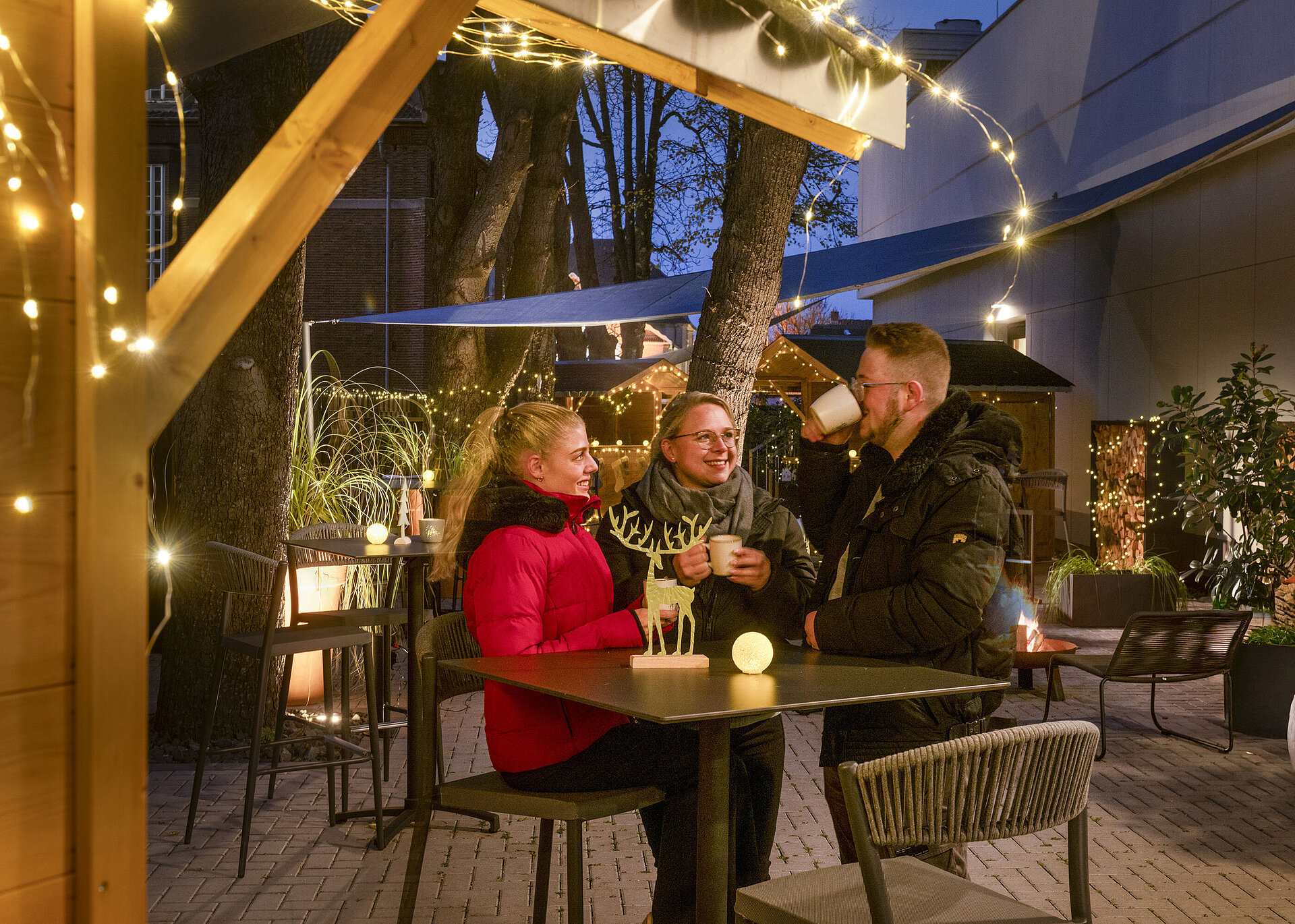 Pueblo Navideño Maritim Tres personas disfrutan de bebidas calientes en el mercado navideño del Hotel Maritim Darmstadt con luces festivas.