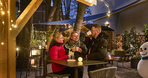 Trois personnes dégustent des boissons chaudes au marché de Noël festif de l'hôtel Maritim avec guirlandes lumineuses.