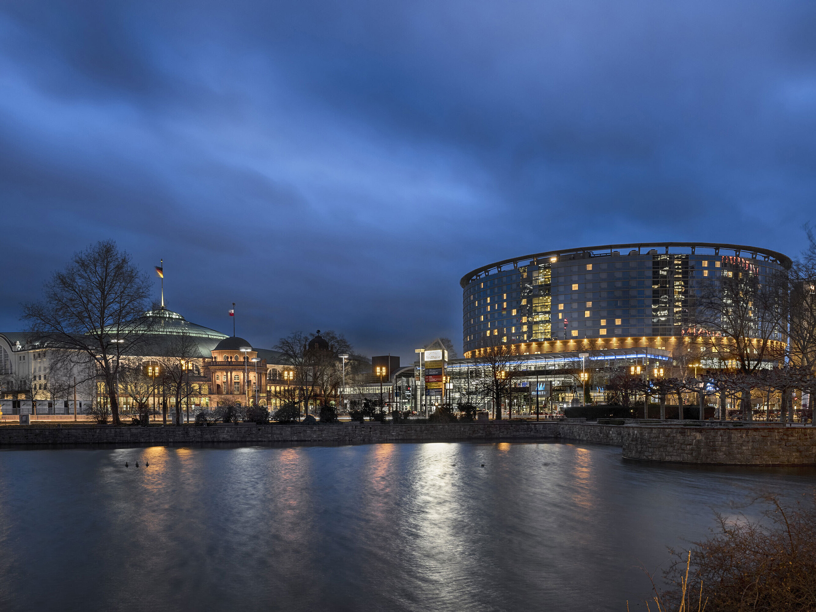 Hôtel Maritim Frankfurt et Festhalle de nuit avec façade en verre illuminée et reflet dans l'étang adjacent.