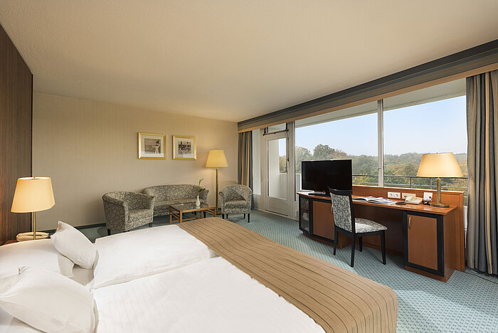 Chambre Comfort Chambre Comfort du Maritim Hotel Kiel avec un coin salon confortable, un bureau, un grand lit et une vue magnifique sur la verdure.