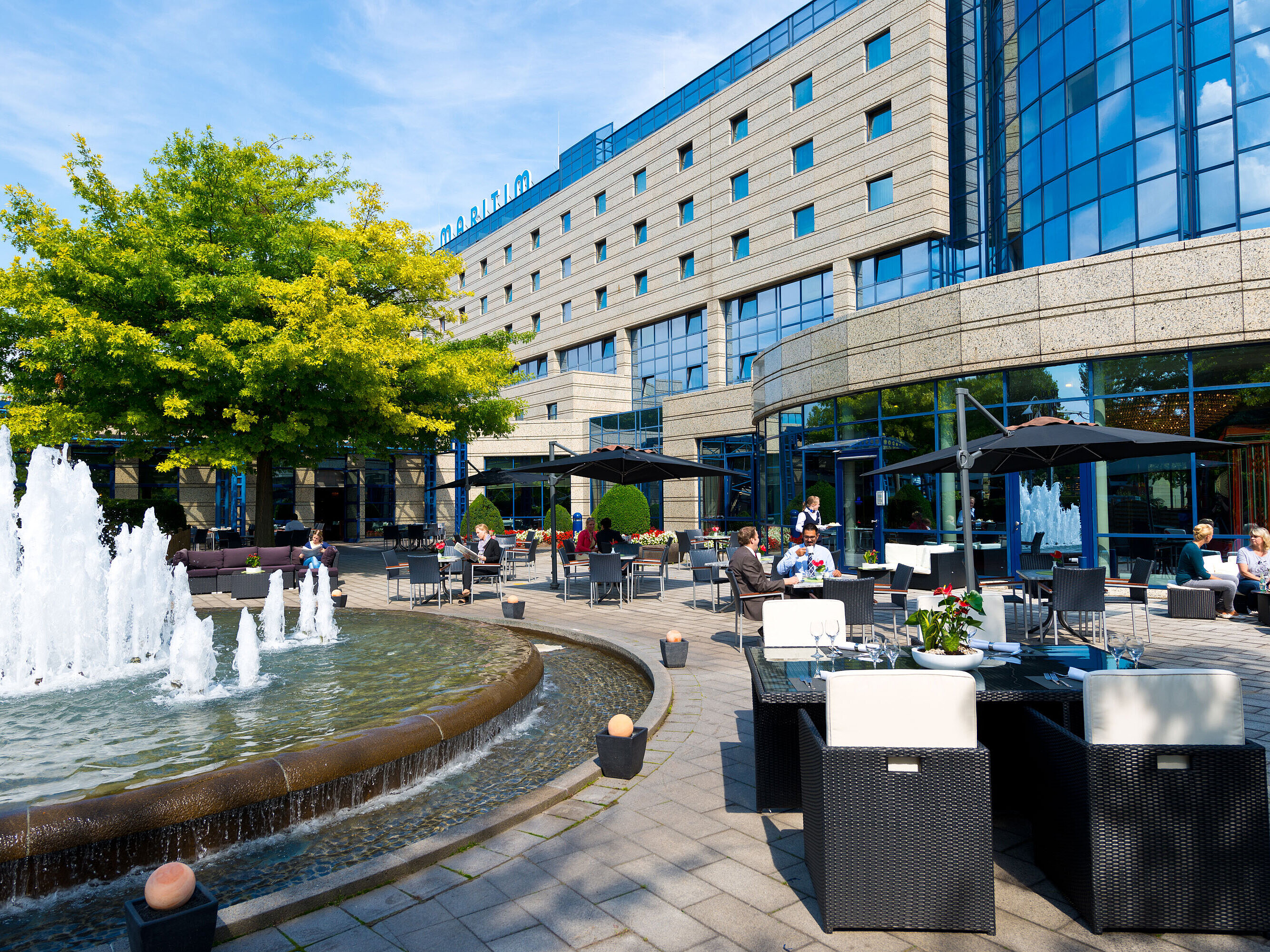 Terrasse de l'hôtel Maritim Bonn avec des coins salon, une fontaine et des clients qui dînent et se détendent en plein air.