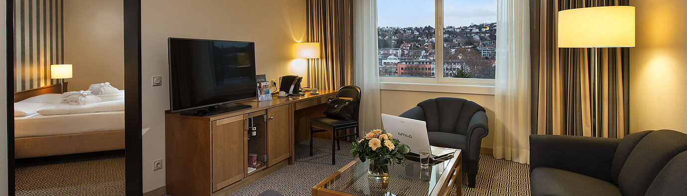 Suite Junior Salon avec canapé, télé, bureau et vue sur la ville dans la suite de l’hôtel Maritim Stuttgart