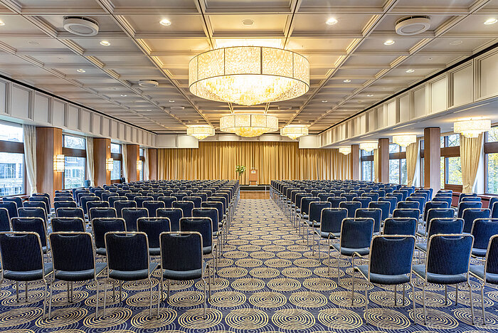 Saal Maritim en el Maritim Hotel Kiel, gran sala de conferencias con asientos en fila, lámparas de araña y un escenario delante de un telón.