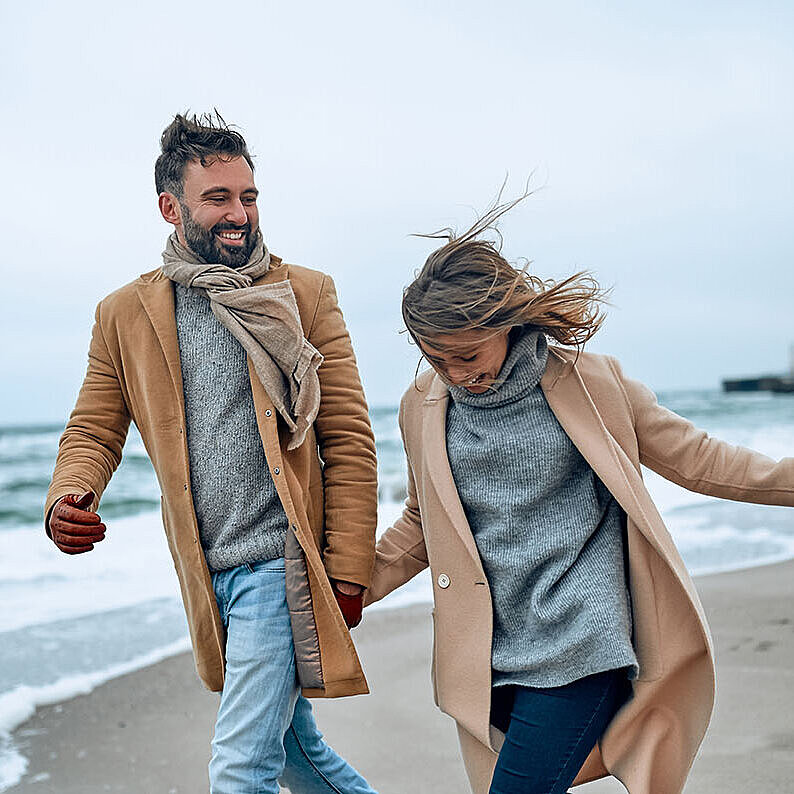 © Valerii Apetroaiei - AdobeStock.com Couple walking along the beach on a windy day, wearing warm clothing.