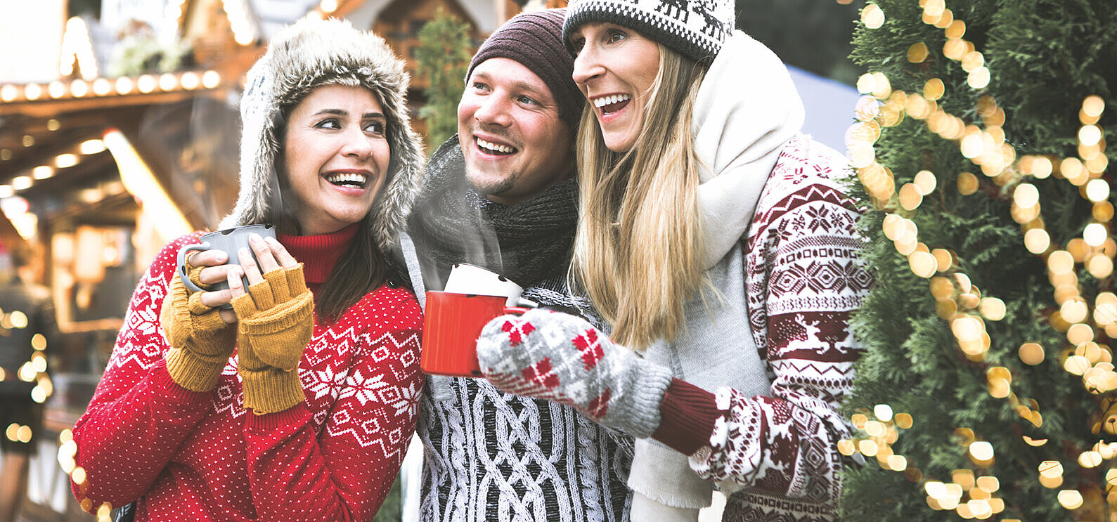 © drubig-photo - Adobestock.com Three laughing friends in winter clothing with hot drinks at a Christmas market