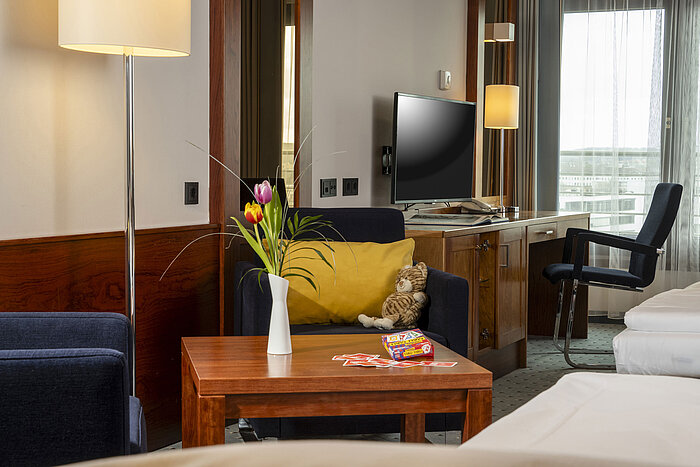 Family room at Maritim Hotel Darmstadt with cozy armchair, table with playing cards and tulip vase, and a desk with TV.