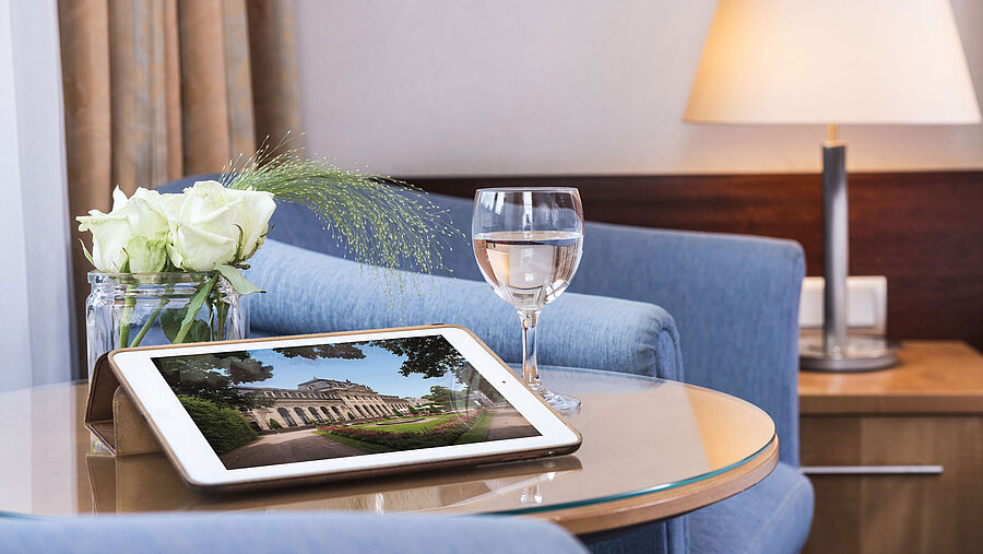 Acogedora zona de descanso en Maritim Hotel Fulda con tablet, vaso de agua y rosas blancas en mesa de vidrio.