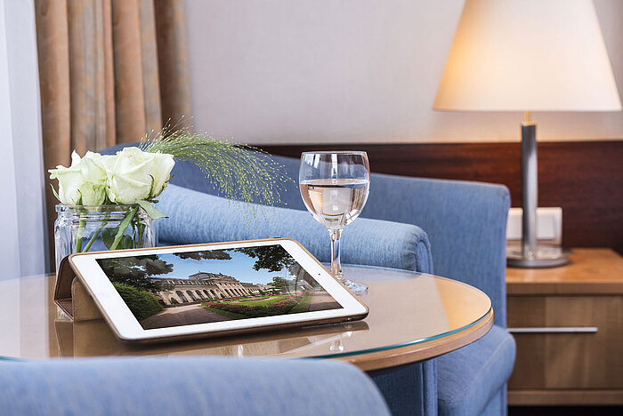Acogedora zona de descanso en Maritim Hotel Fulda con tablet, vaso de agua y rosas blancas en mesa de vidrio.