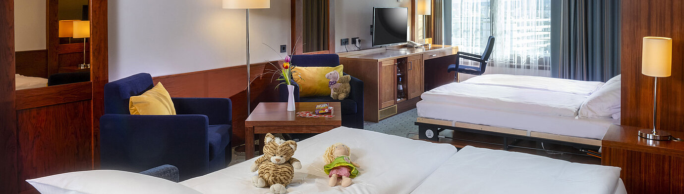 Camera familiare al Maritim Hotel Darmstadt con giocattoli sul letto, scrivania con TV e area salotto accogliente.