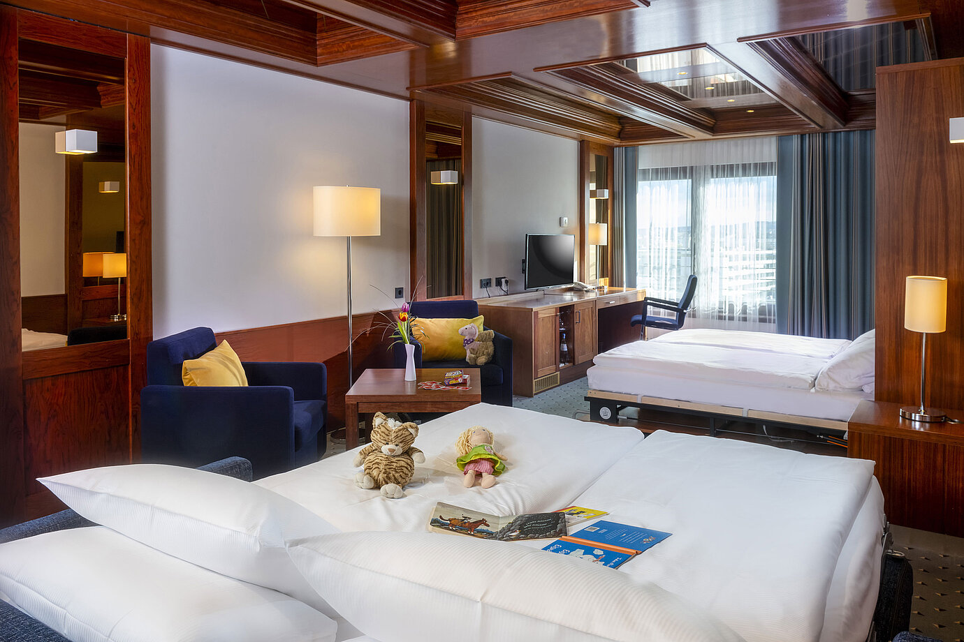 Family room at Maritim Hotel Darmstadt with toys on the bed, a desk with TV, and a cozy seating area.