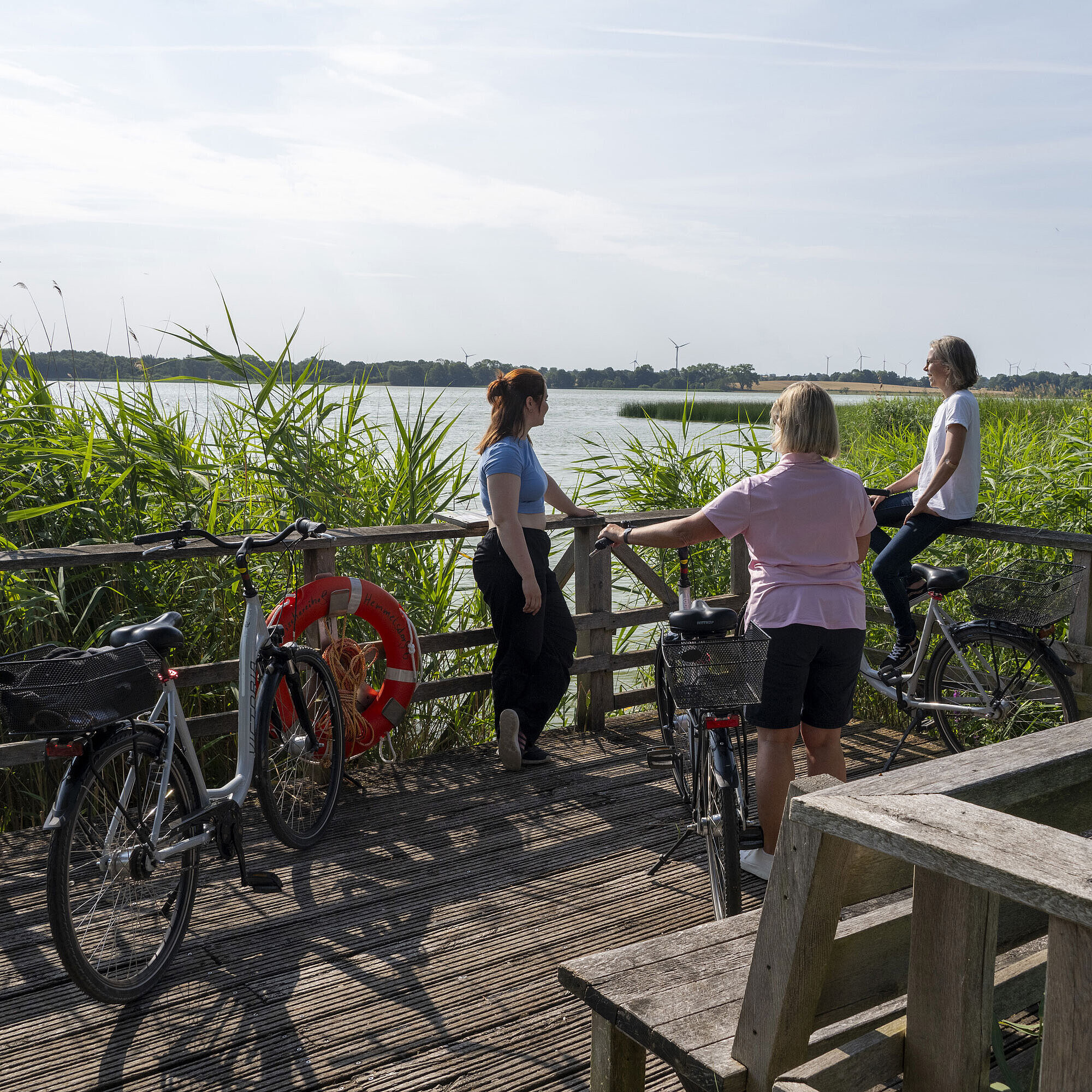 Tres mujeres descansan durante un paseo en bici en un muelle con vista al lago en Maritim Seehotel Timmendorfer Strand.