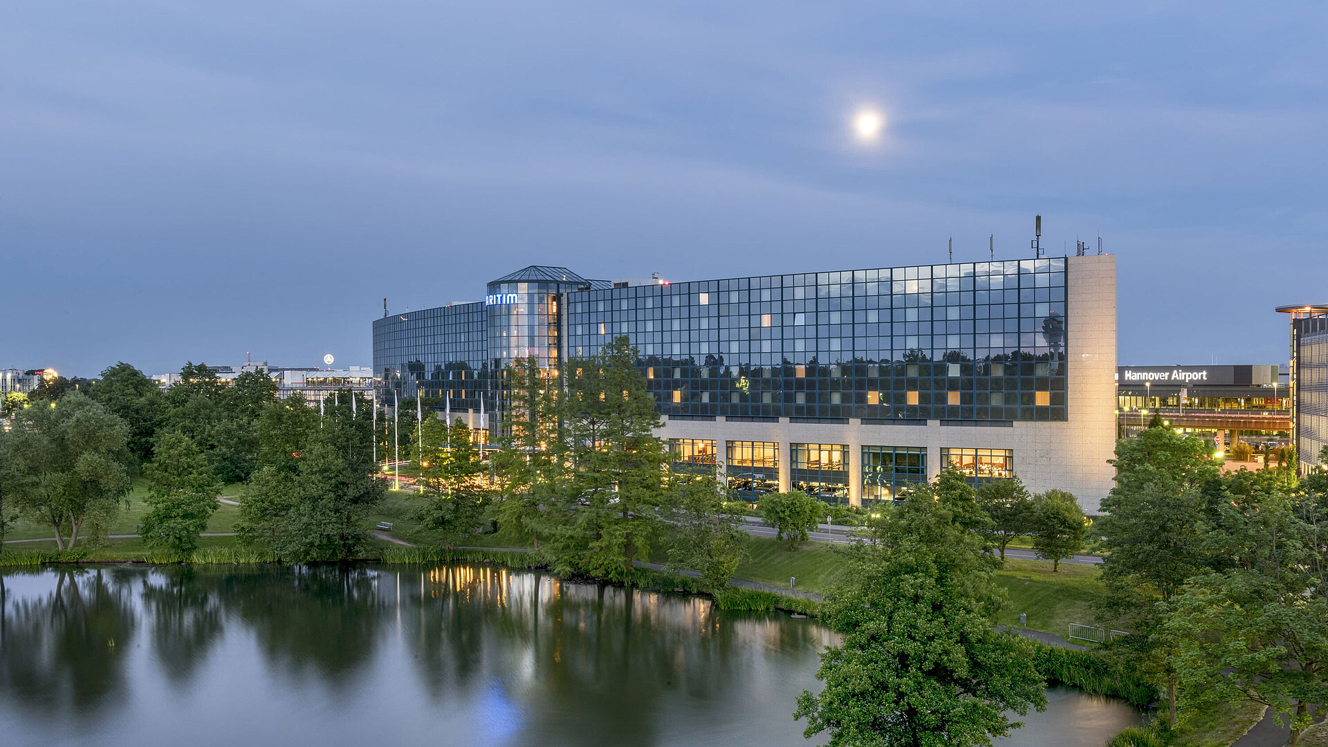Illuminated Maritim Hotel Hannover with a glass facade and lake reflection at dusk.