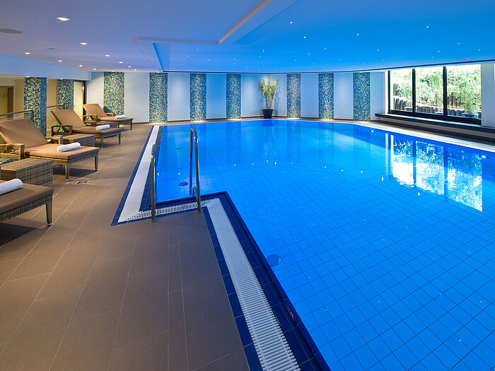 Bright and modern indoor pool at Maritim Hotel Darmstadt with lounge chairs and large windows overlooking greenery.