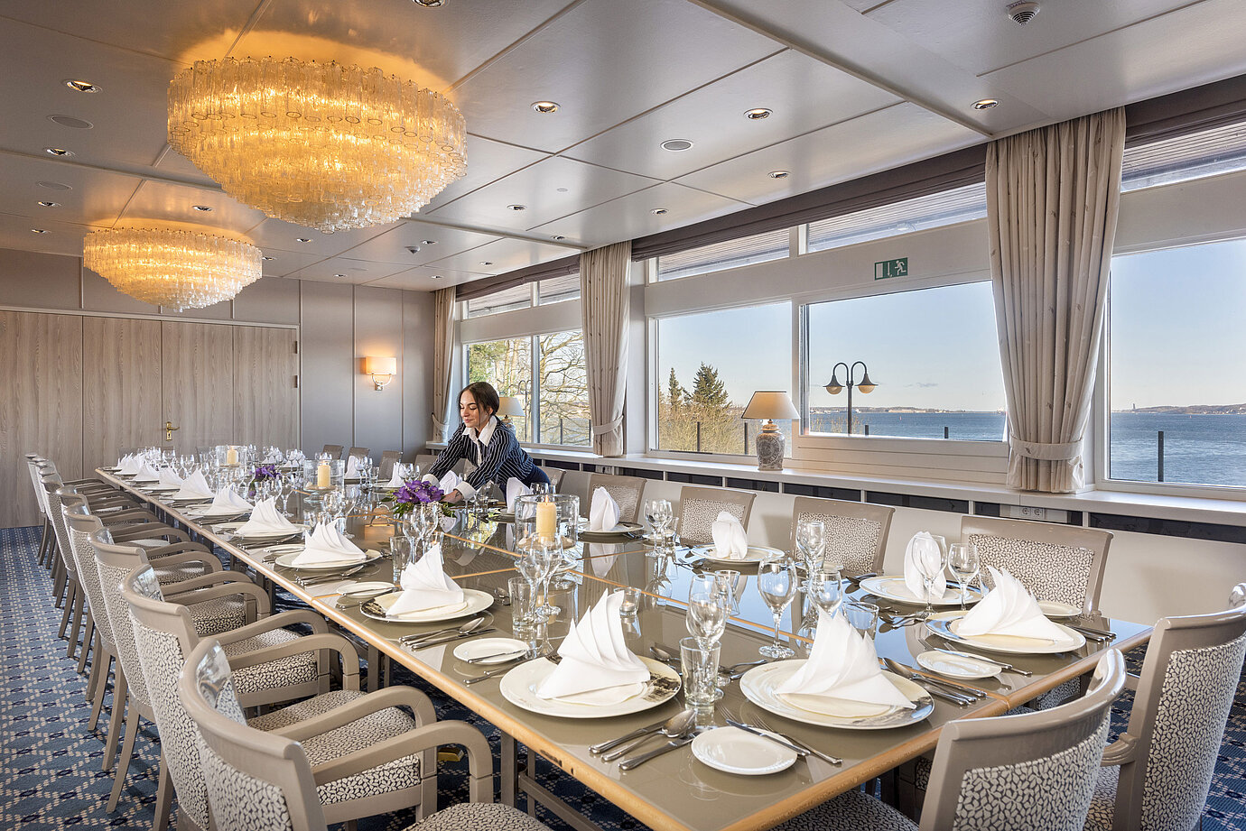 Salón Oslo en el Maritim Hotel Kiel, larga mesa festiva con vistas al mar, lámpara de araña, personal arreglando la sala.