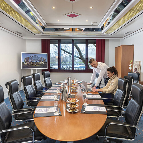 Dos personas trabajan en el Salon Boardroom del Maritim Colonia con portátiles en una mesa de conferencia con bebidas y snacks