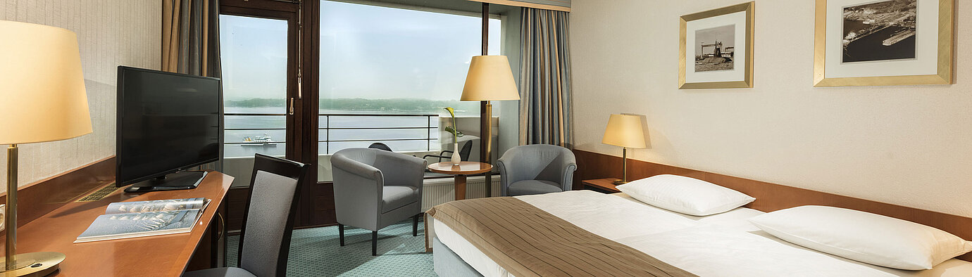 Habitación superior del Maritim Hotel Kiel con escritorio, zona de estar, cama doble grande y balcón con vistas al fiordo.