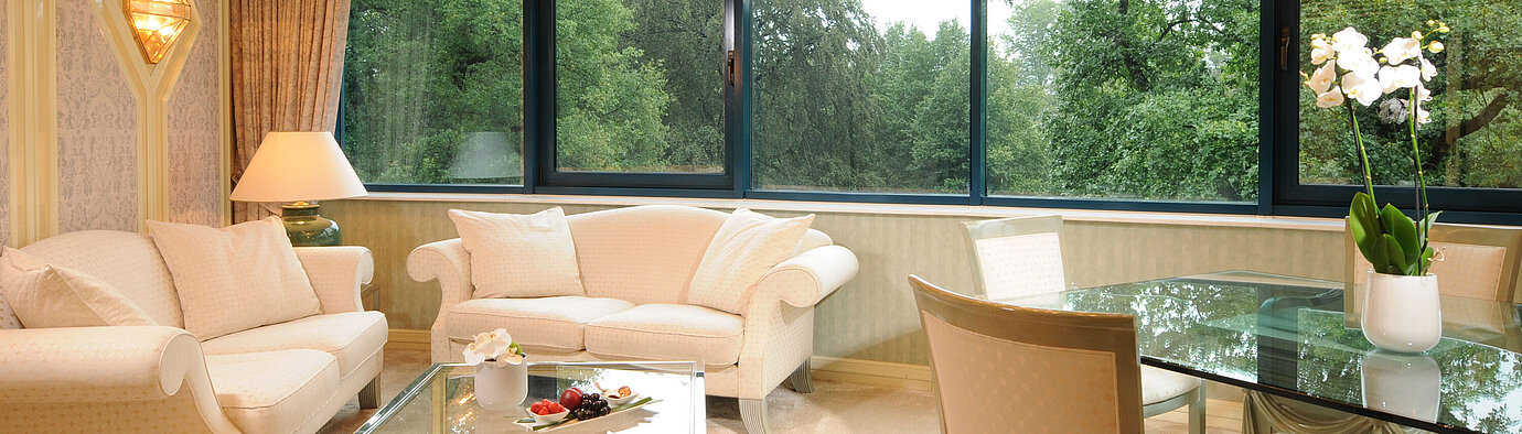 Suite luxueuse avec coin salon élégant, table en verre et grande fenêtre panoramique avec vue verte.